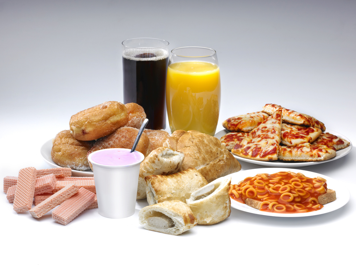 A selection of processed food and drink on a white background including pIzza, cola, buscuits and sausage rolls